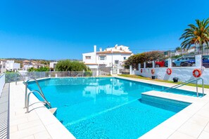 A swimming pool with a clear blue water, surrounded by a white fence, and a view of a white building with a red car parked in front.