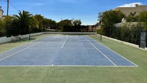 Cancha de tenis exterior con red, rodeada de vegetación y edificios.