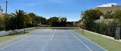An outdoor tennis court with a net, surrounded by greenery and buildings.