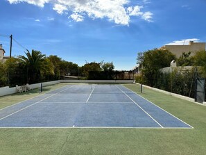 An outdoor tennis court with a net, surrounded by greenery and buildings.