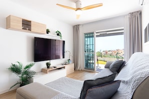 Ein modernes Wohnzimmer mit einem Flachbildfernseher, Holzregalen, einem Deckenventilator und einer Schiebetür zum Balkon.