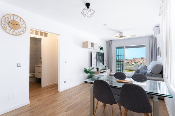 A modern living room with a glass dining table, grey chairs, a sofa, and a view of a cityscape through large windows.