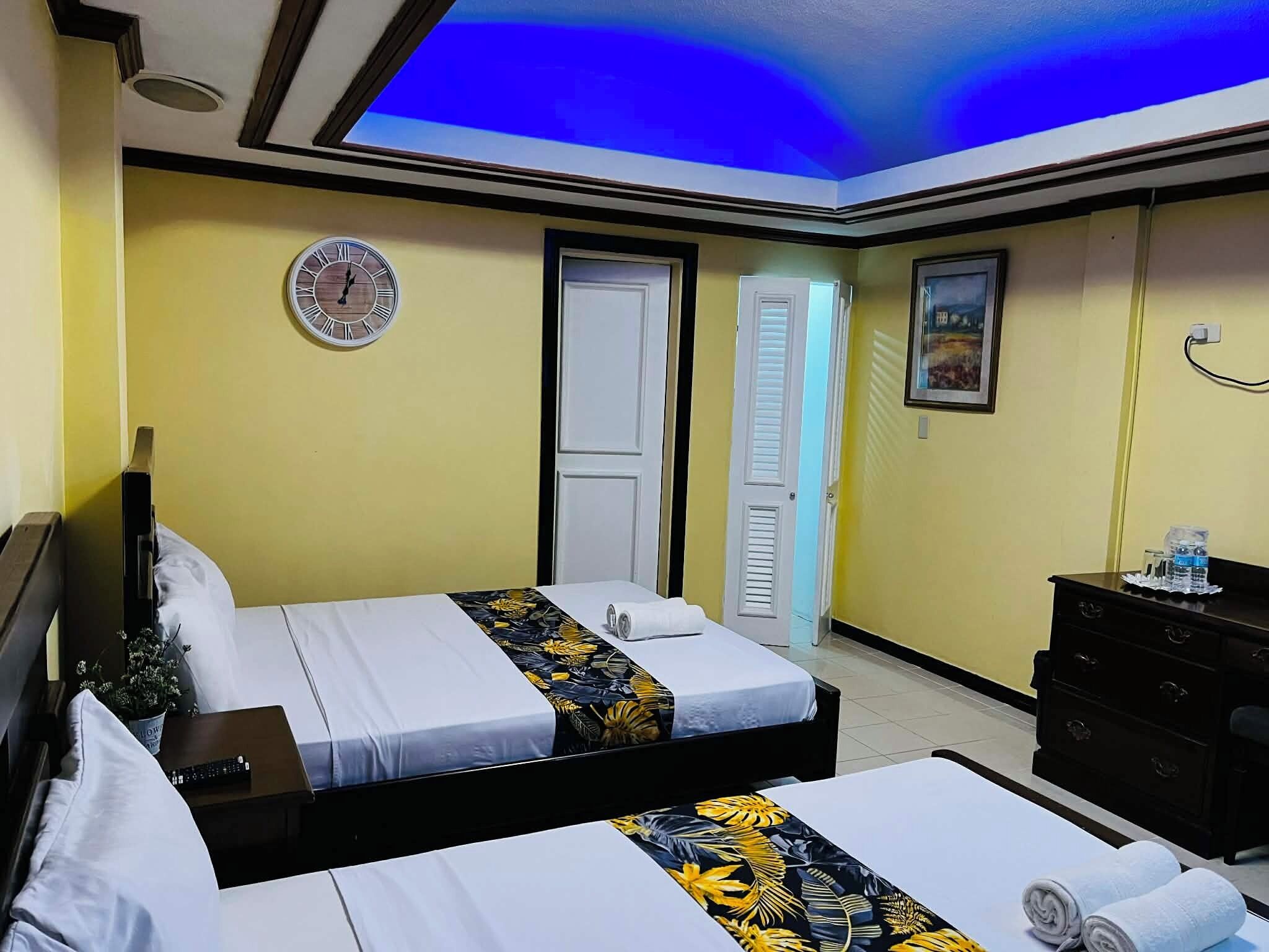 A hotel room with two beds, a ceiling light, a clock, a door, a dresser, and a painting.