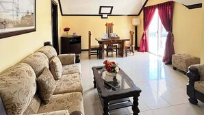A spacious living room with a sofa set, a coffee table, a dining table with chairs, and a large window with curtains.