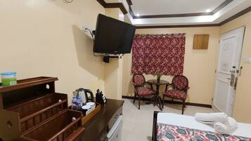 A hotel room with a bed, a TV mounted on the wall, a small table with chairs, a wooden dresser, and a window with a floral curtain.