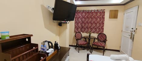 Une chambre d’hôtel avec un lit, un téléviseur fixé au mur, une petite table avec des chaises, une commode en bois et une fenêtre agrémentée d’un rideau à motifs floraux.