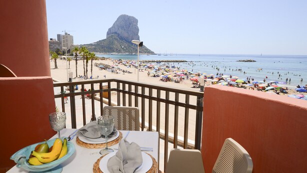 Un balcón con mesa lista para comer, con vista a una playa concurrida, palmeras y una formación rocosa distintiva al fondo.