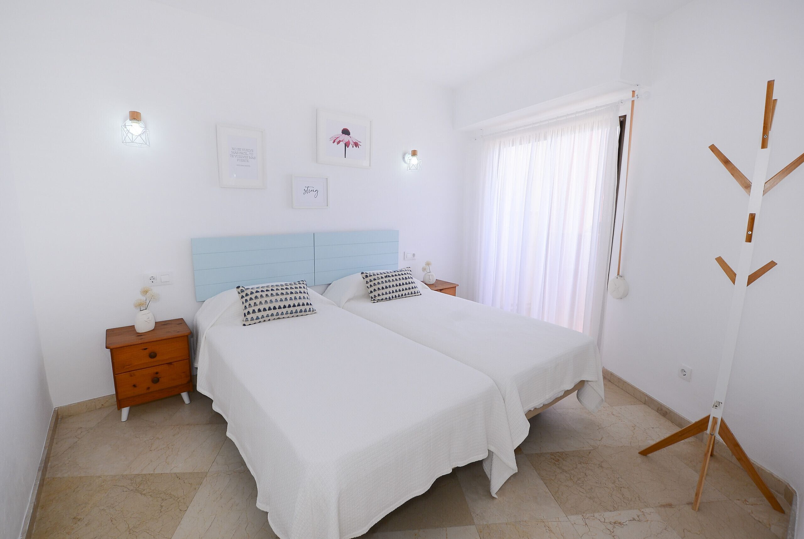 A bedroom with two beds, a nightstand, a wall-mounted light, and a window with curtains.