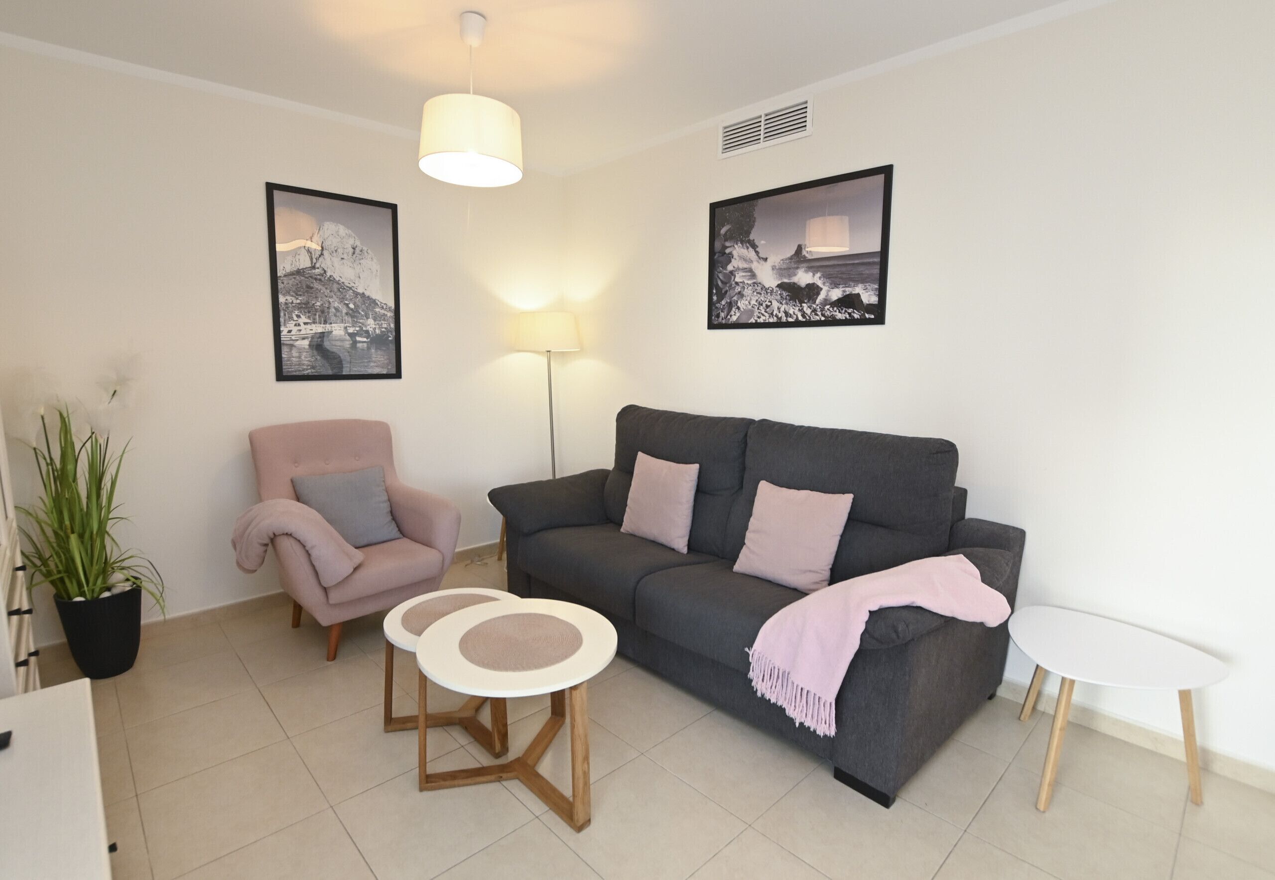 A living room with a grey sofa, a pink armchair, a small white table, and a floor lamp. There are two framed pictures on the wall.