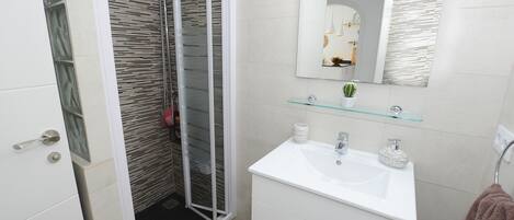 A bathroom with a glass shower enclosure, a white sink with a glass shelf, and a mirror with a textured wall backsplash.