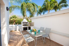 An outdoor patio with a barbecue grill, a dining table with chairs, and a white wall.