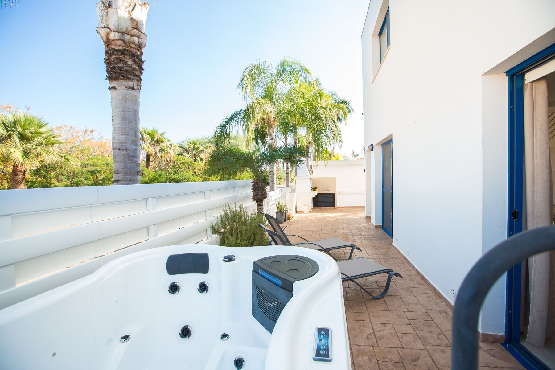 A hot tub in a covered outdoor area with a patio chair and a small table, surrounded by palm trees and a white building with a blue door.