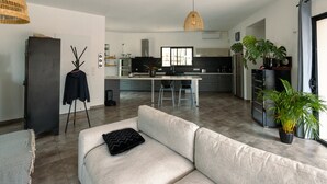 Un salon moderne avec un canapé, un coin repas comprenant une table et des chaises, une cuisine en arrière-plan et une grande fenêtre laissant entrer la lumière naturelle.