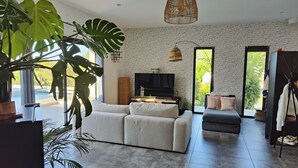 Un salon moderne avec un grand canapé, un téléviseur et un coin repas. De grandes fenêtres offrent une vue sur la verdure environnante.