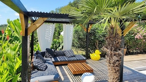Un espace extérieur aménagé, abrité et doté de bancs en bois et de coussins, entouré d’une végétation luxuriante et d’un palmier.