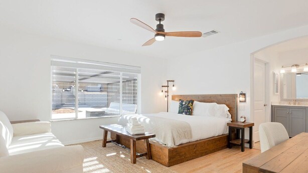A bedroom with a bed, bedside tables, a ceiling fan, a window, and a view of the outside. - Of Wine & Gods - Hot Tub, BBQ and Fire Pit (Joshua Tree)