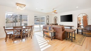 A spacious living room with a wooden dining table, a leather sofa, and a flat-screen TV mounted on the wall. - Of Wine & Gods - Hot Tub, BBQ and Fire Pit (Joshua Tree)