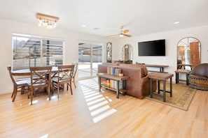 A spacious living room with a wooden dining table, a leather sofa, and a flat-screen TV mounted on the wall. - Of Wine & Gods - Hot Tub, BBQ and Fire Pit (Joshua Tree)