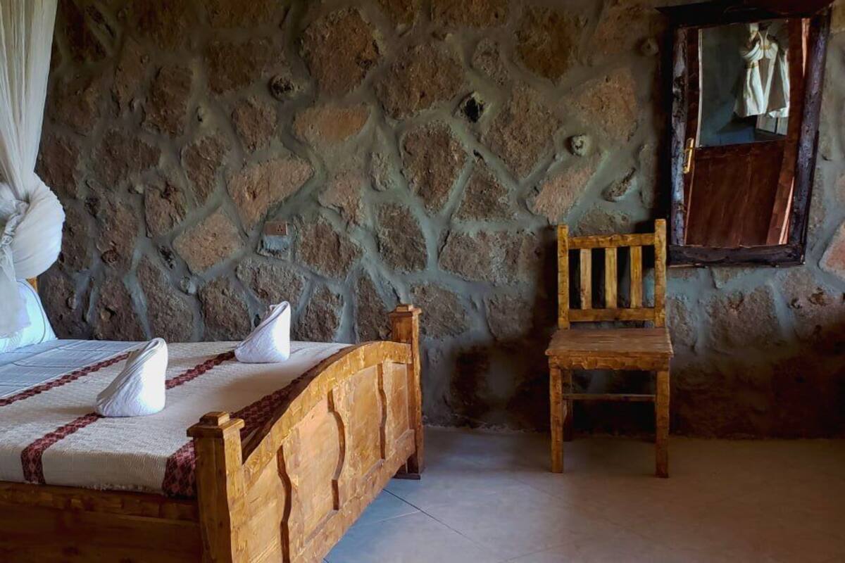 A bed with a wooden headboard, a wooden chair, and a stone wall.