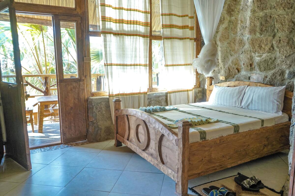 A wooden bed with a headboard, a stone wall, and a view of palm trees outside.