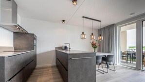 Una cocina moderna con una isla central, un comedor con sillas, luces colgantes y vistas al exterior.