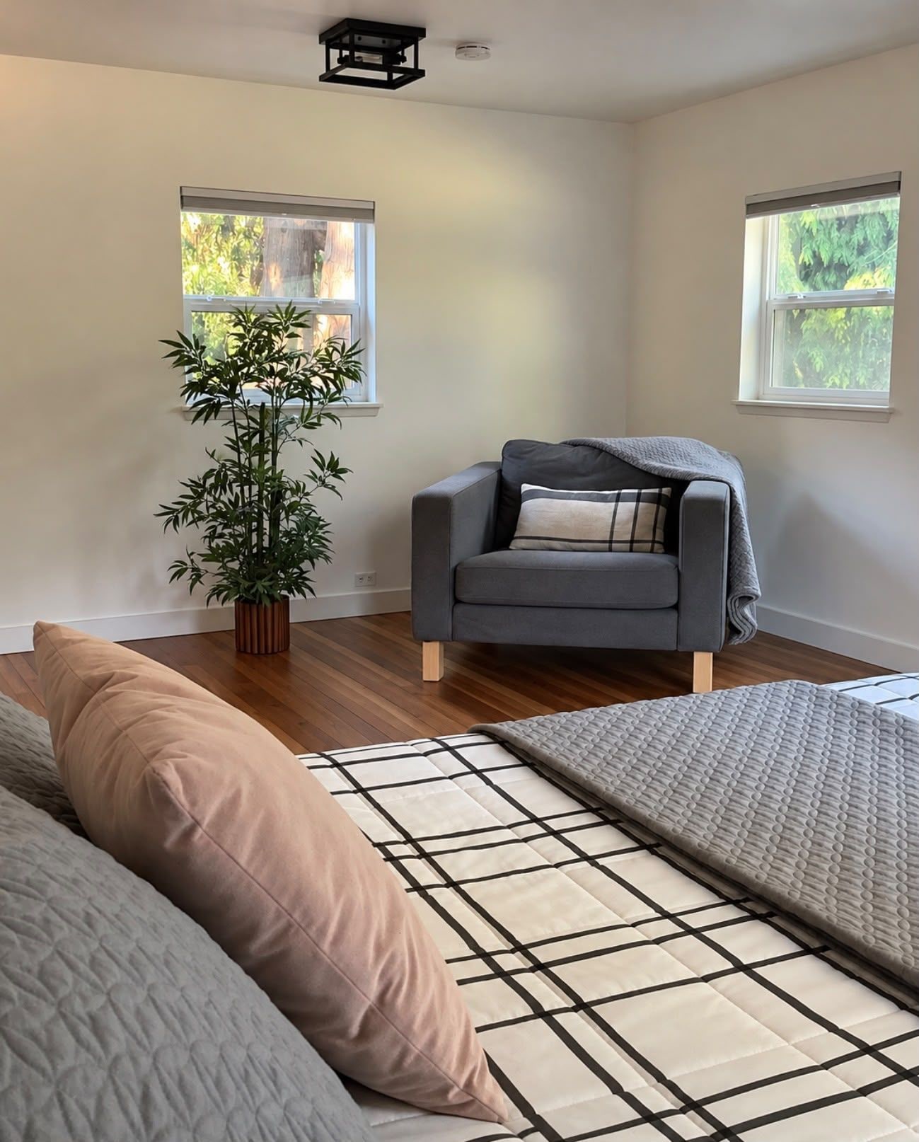 Ein Zimmer mit grauem Sofa, einer großen Topfpflanze, einem gemusterten Teppich und zwei Fenstern mit Blick auf Grünflächen.