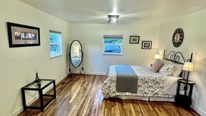 Un dormitorio con suelo de madera, una cama con ropa de cama floral, dos mesitas de noche con lámparas, un ventilador de techo y cuadros en las paredes.