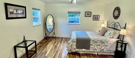 A bedroom with a wooden floor, a bed with floral bedding, two bedside tables with lamps, a ceiling fan, and framed pictures on the walls.
