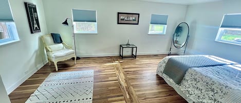 A room with wooden flooring, a bed with floral bedding, a chair, a small table, and a mirror.