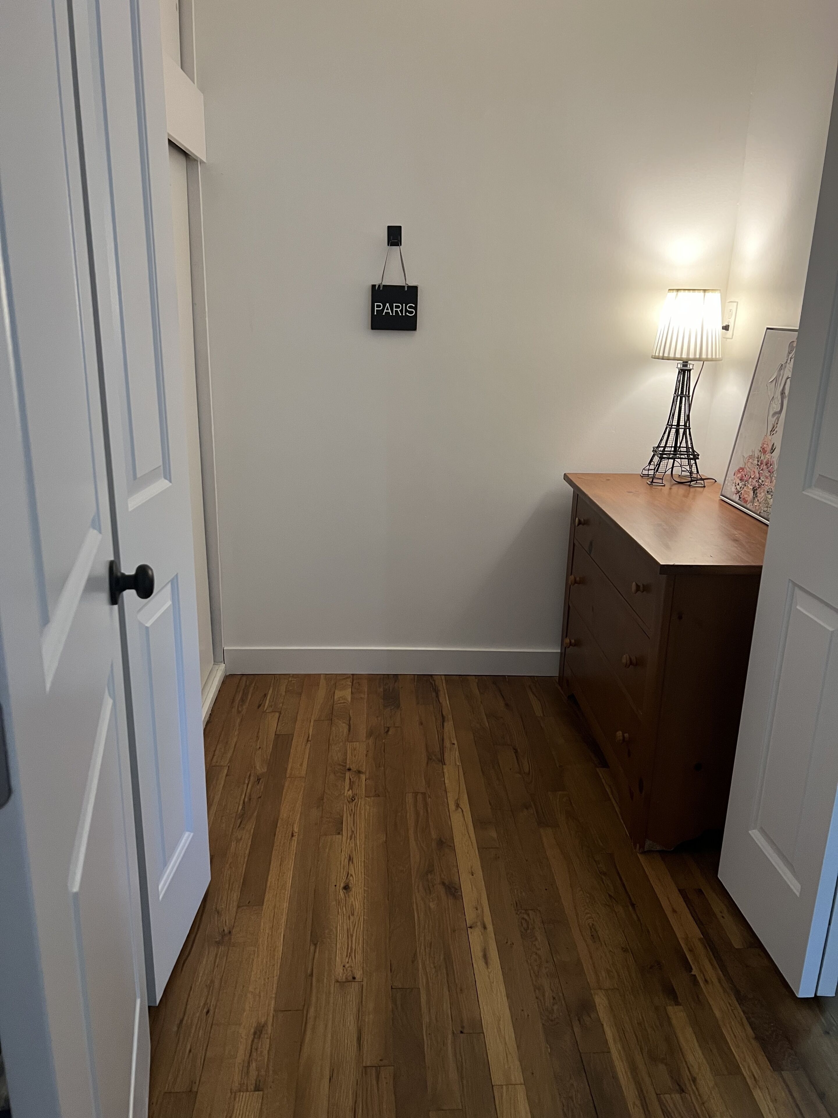 Una habitación con piso de madera, un escritorio con cajones, una lámpara y un cartel con la palabra 'PARIS' en la pared.
