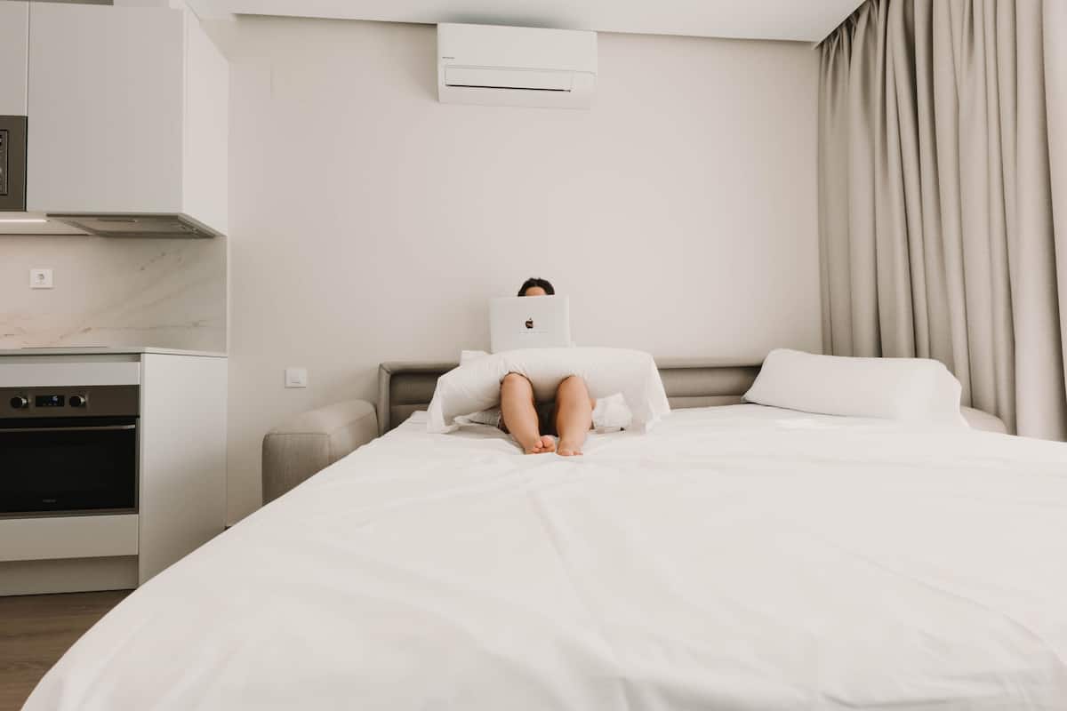 A modern hotel room with a bed, a small bedside table, a built-in oven, and a wall-mounted air conditioning unit.