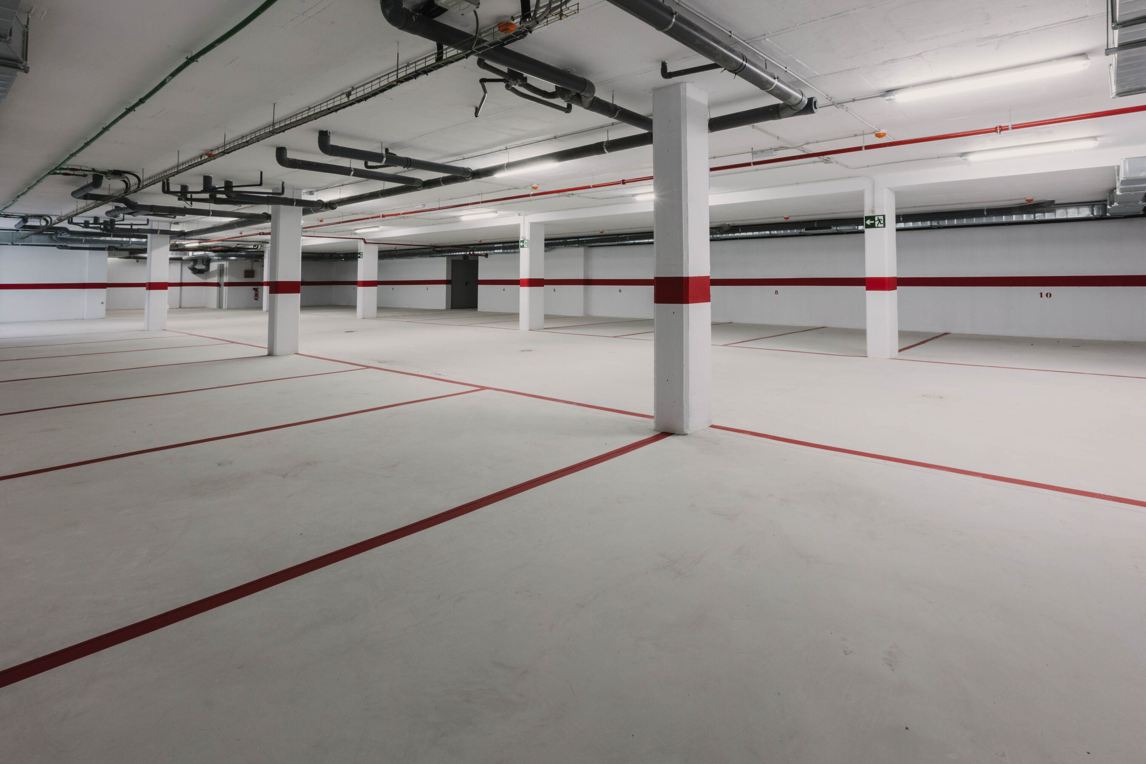 An empty parking garage with white floors and red lines, white pillars with red stripes, and visible ceiling pipes.