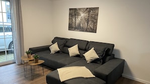 Uma sala de estar moderna com um sofá seccional cinza escuro, duas mesas laterais com plantas, um cobertor bege e uma obra de arte com tema de floresta na parede.