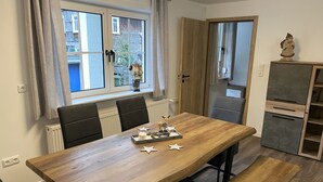 Uma área de refeições com uma mesa de madeira, cadeiras pretas e um banco. Há uma janela com cortinas, um radiador e uma porta que dá para o exterior.