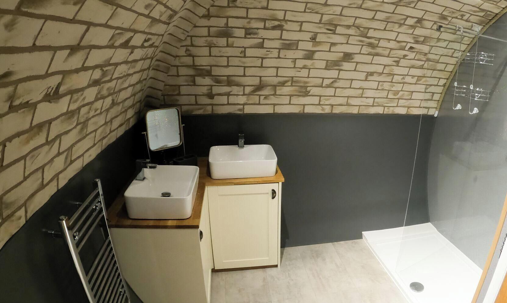 Un bagno con parete in mattoni, un piccolo mobile con specchio, due lavandini e una zona doccia.