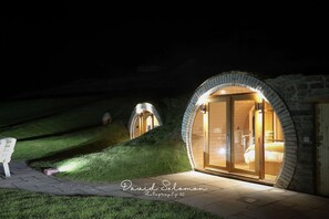 Twee gebogen, stenen gebouwen met glazen deuren, verlicht in de nacht, gelegen op een grashelling.