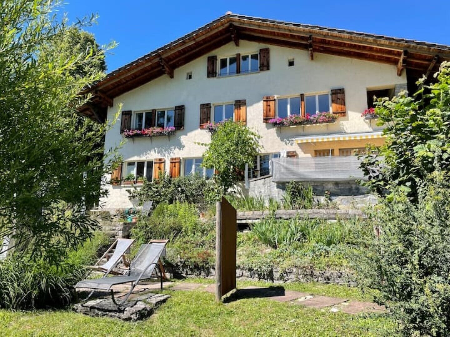 A two-story building with a sloped roof, multiple windows with shutters, and a balcony with flower boxes. There are two lounge chairs in the foreground.