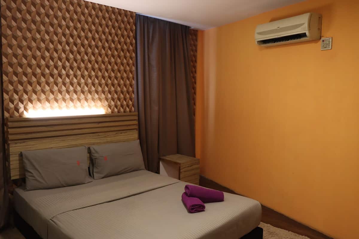A hotel room with a bed, a headboard with a geometric pattern, an air conditioning unit, and a small wooden side table.