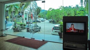 Um hall de hotel com uma grande porta de vidro que dá para a rua. No interior, há uma tela de televisão exibindo um programa.