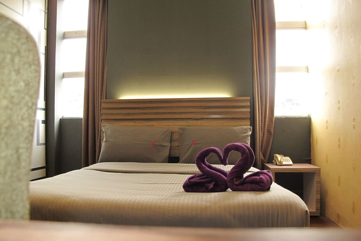 A hotel room with a bed, two pillows, a nightstand with a phone, and a window with curtains.
