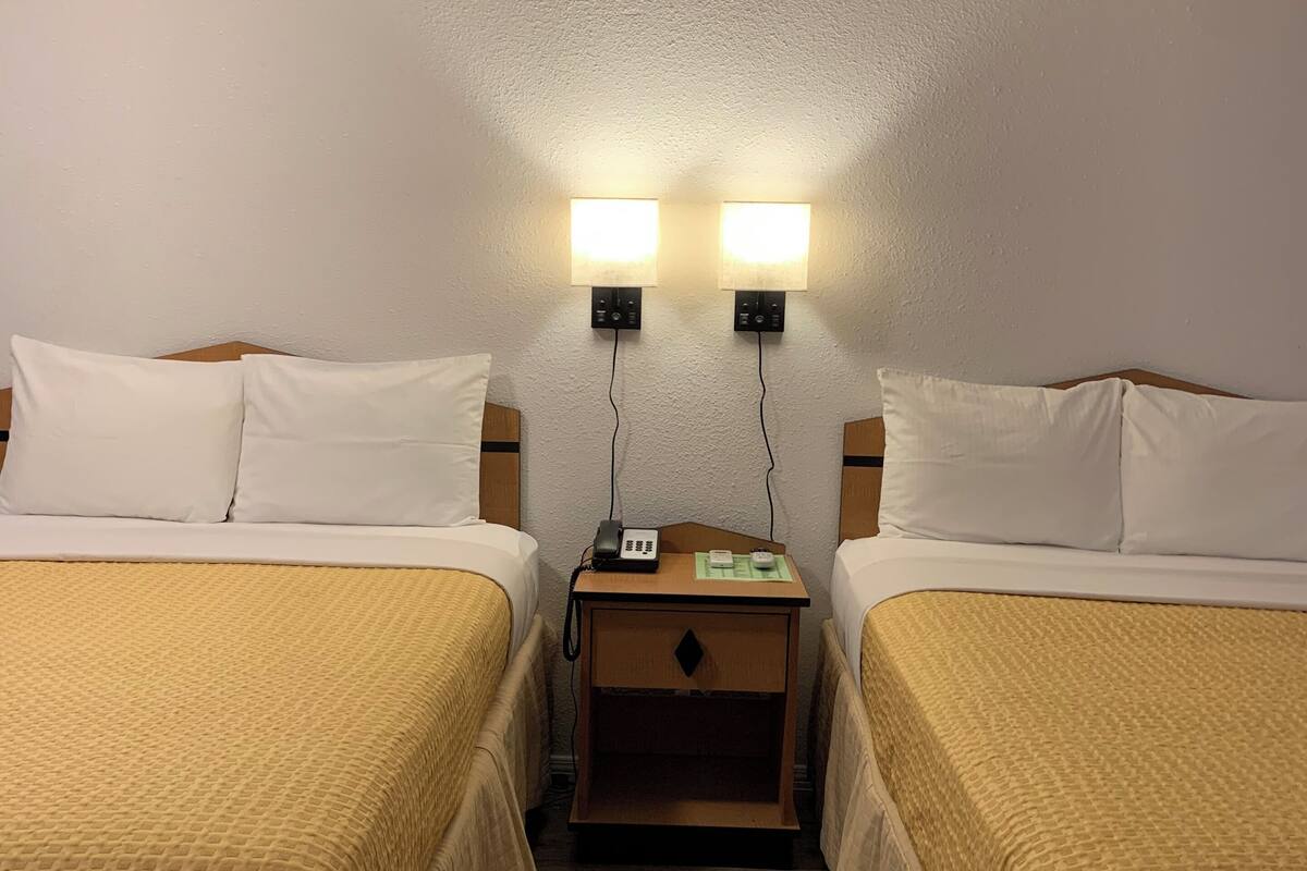Habitación de hotel con dos camas individuales, cada una con cabecero de madera, una mesita de noche de madera entre las camas y dos luces fijadas a la pared.