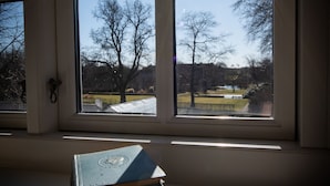 Une fenêtre donnant sur des arbres et un plan d’eau, une pile de livres posée sur un rebord, et la lumière du soleil qui filtre à travers la fenêtre.