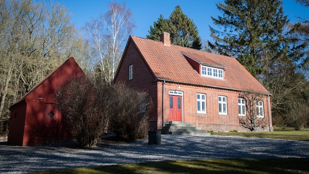 Une maison en briques rouges, avec une porte rouge et des fenêtres blanches, entourée d’arbres et bordée d’une allée en gravier.