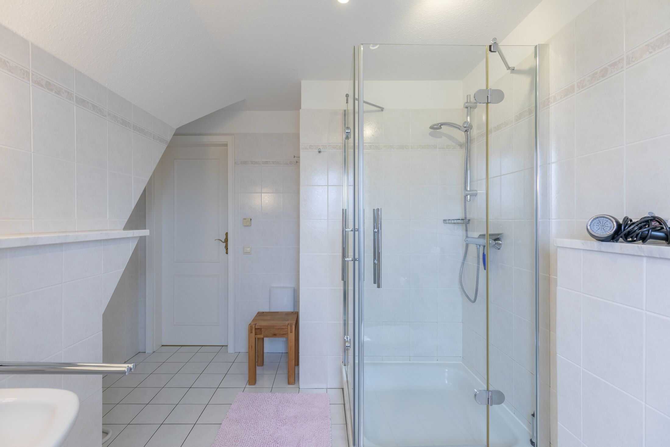 Ein modernes Badezimmer mit einer gläsernen Duschkabine, einem hölzernen Hocker und einem weißen Fliesenboden.