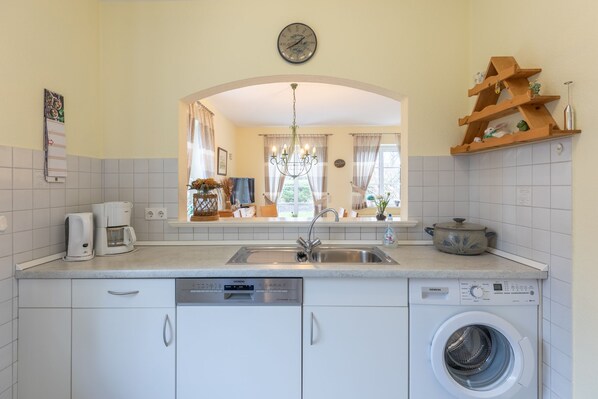 A kitchen with white cabinets, a dishwasher, a washing machine, a coffee maker, a sink, and a wooden shelf with decorative items.