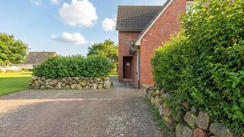 Een bakstenen gebouw met een verharde oprit die naar een deur leidt, omgeven door groen en een stenen muur.