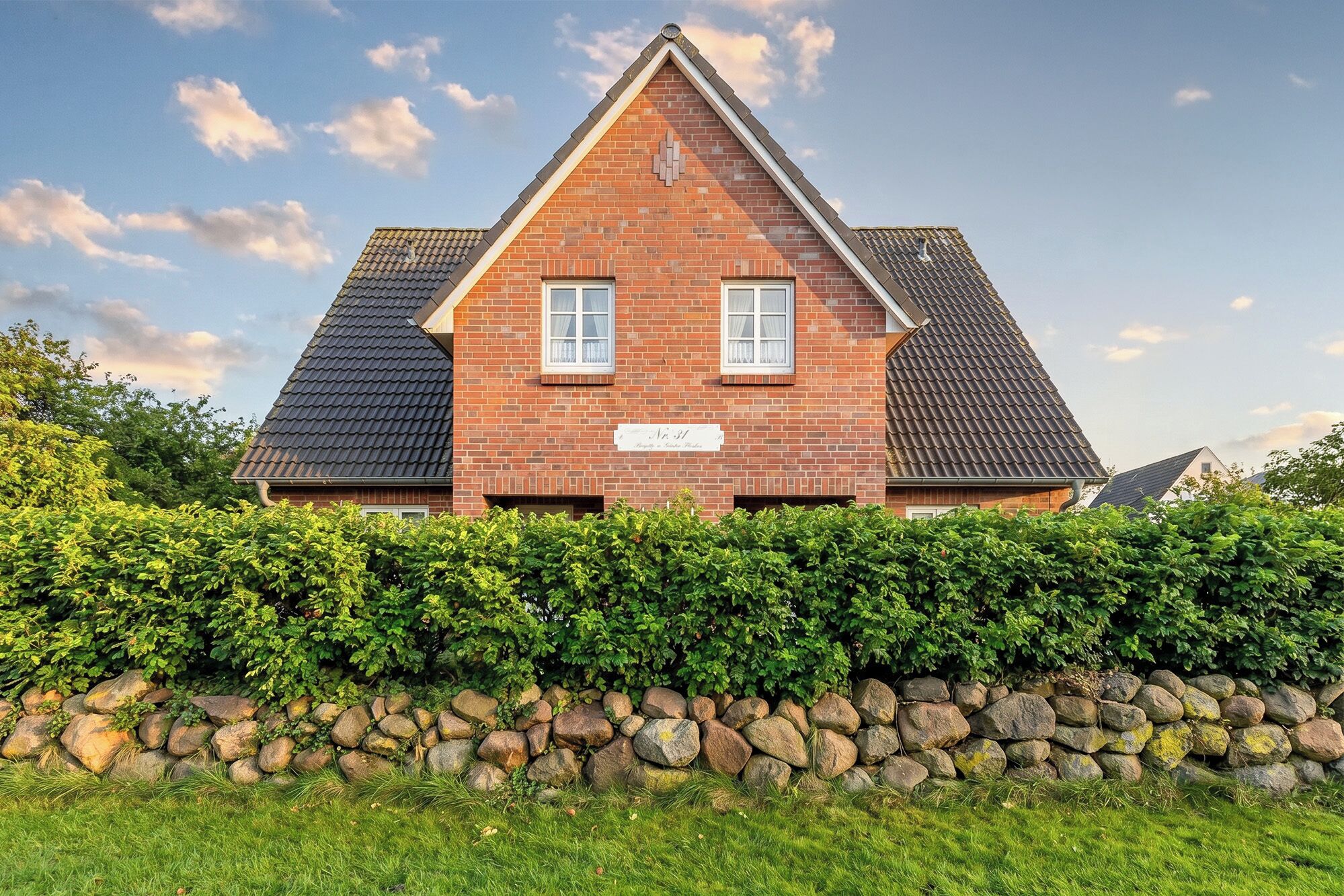 Een tweelaags huis met een bakstenen gevel, twee ramen met witte omlijstingen, en een stenen muur ervoor.