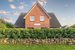 A two-story house with a brick facade, two windows with white frames, and a stone wall in front.