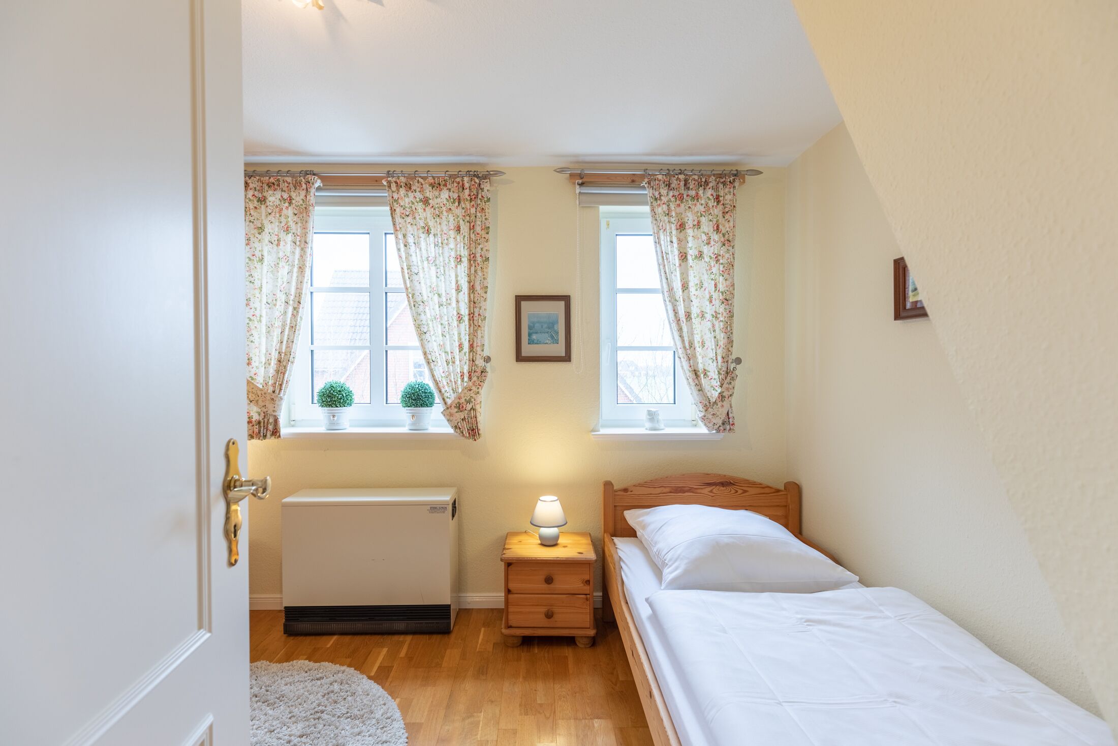 A bedroom with a wooden bed, a nightstand with a lamp, a radiator, and two windows with floral curtains.