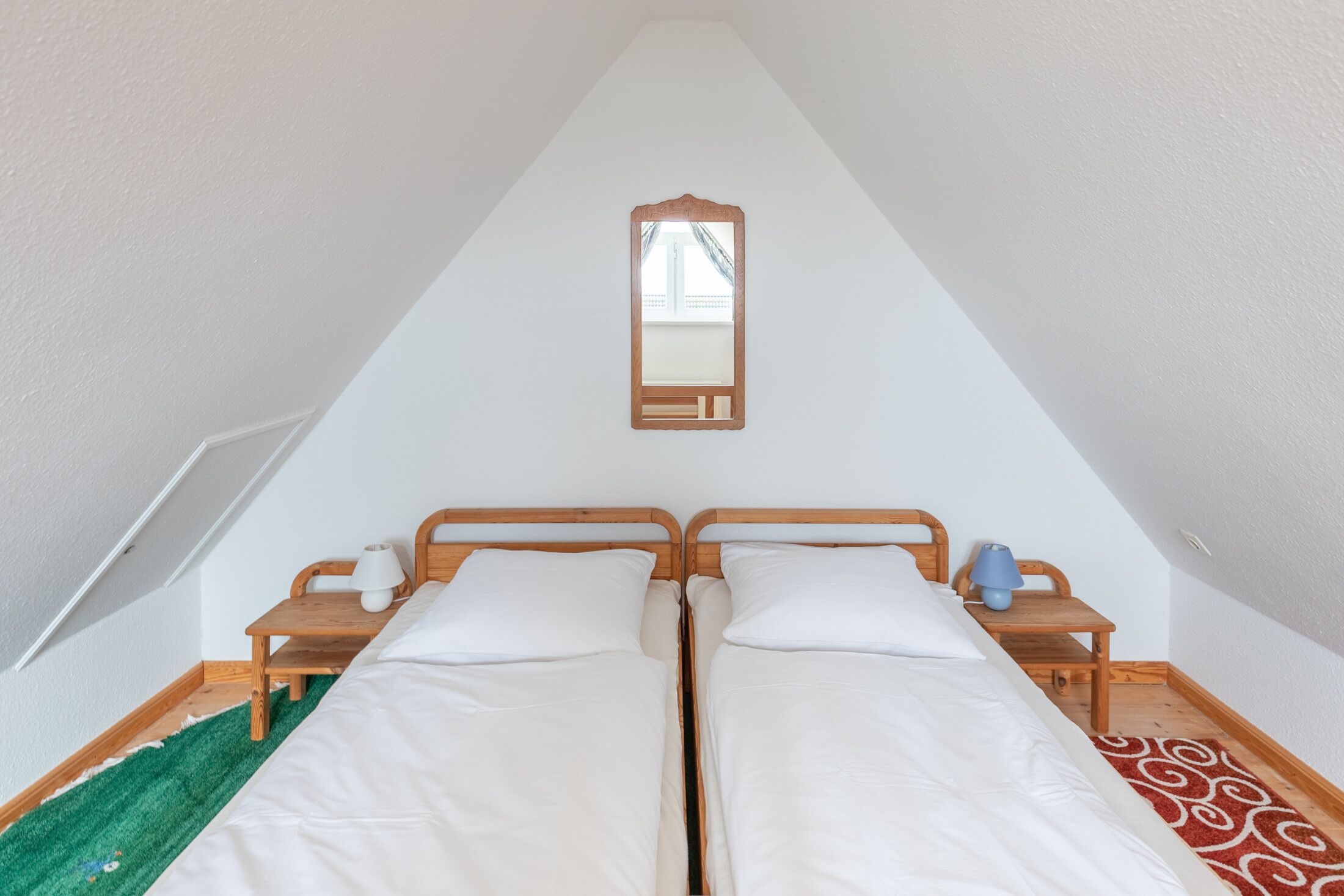 A room with two beds, a wooden headboard, a small table with a lamp, and a mirror on the wall.